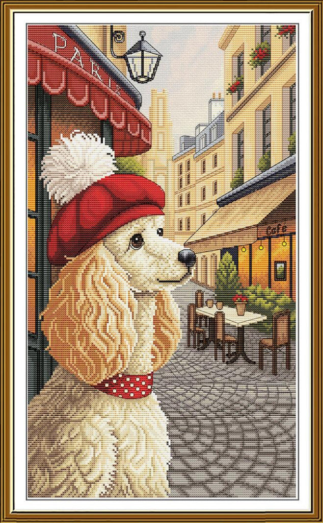 A framed needlepoint or cross-stitch style artwork showing a cream-colored poodle wearing a red beret with a white pom-pom and a red polka-dot collar, set against a Parisian street scene with café tables and buildings.
