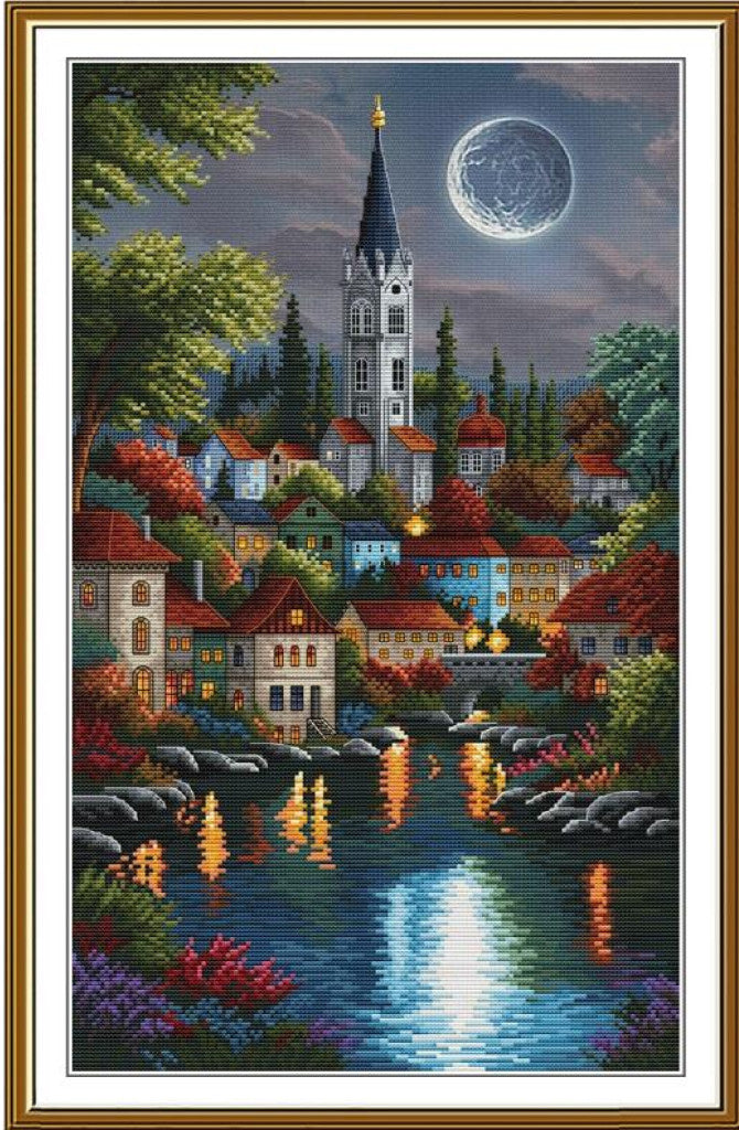 A framed needlepoint canvas featuring a picturesque European village at night under a full moon, with a church spire, colorful houses, trees, and reflections in a river.