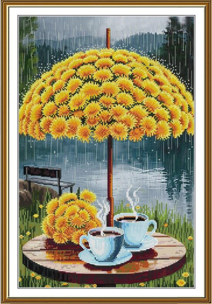 A framed needlepoint or cross-stitch style artwork showing a large yellow flower umbrella over two steaming cups on a round table, with a lake, bench, and rain in the background.