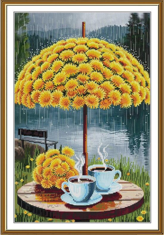 A framed needlepoint or cross-stitch style artwork showing a large yellow flower umbrella over two steaming cups on a round table, with a lake, bench, and rain in the background.