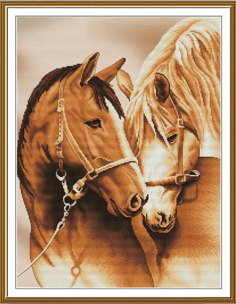 A framed cross-stitch or needlework-style artwork showing two horses in warm tan, brown, cream, and beige tones, with a gold-toned frame and white mat border.