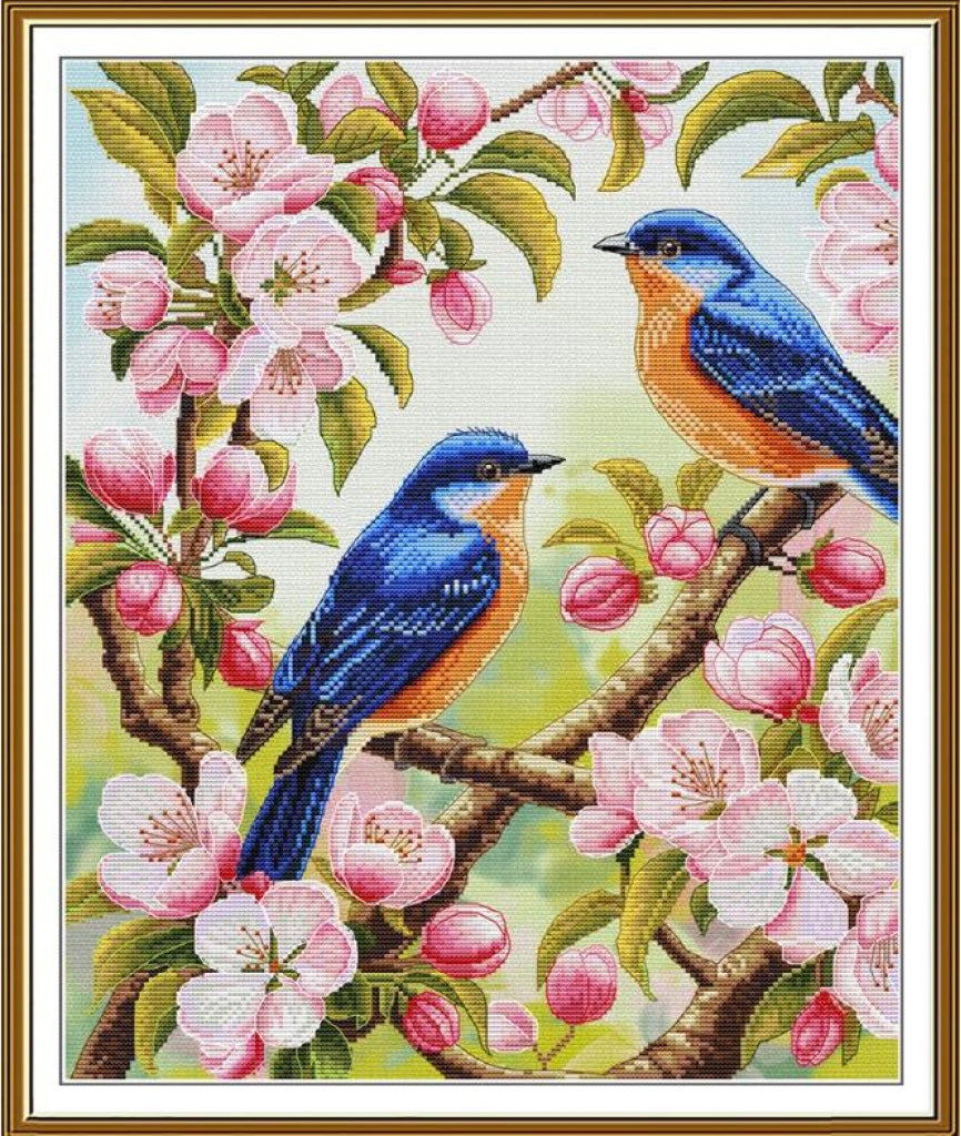 A framed cross-stitch or needlework-style floral and bird artwork showing two blue birds perched on branches with pink blossoms and green leaves, enclosed in a gold-toned frame.