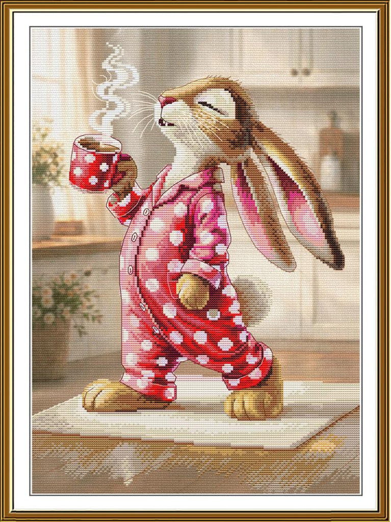 A framed needlepoint or cross-stitch style artwork showing a cartoon rabbit in pink-and-white polka dot pajamas holding a red mug with steam rising, set in a softly lit kitchen scene.