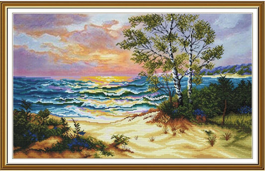 A framed landscape painting depicting a beach scene at sunset, with blue ocean waves, sandy shore, green trees, and a pink and yellow sky, enclosed in a gold-toned frame.