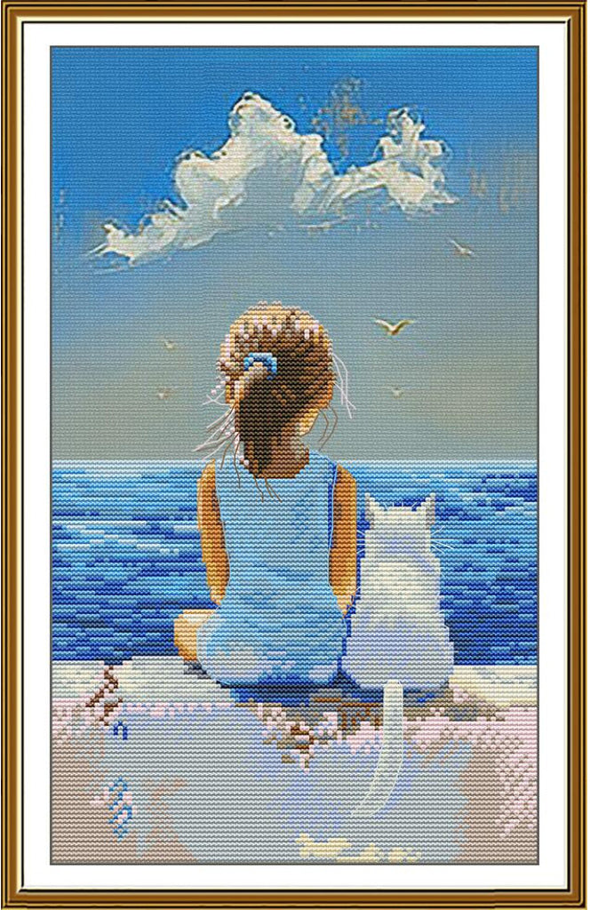 A framed needlepoint canvas featuring a girl with a ponytail sitting by the ocean next to a white cat, under a blue sky with clouds. The artwork is enclosed in a white mat and a gold-colored frame.