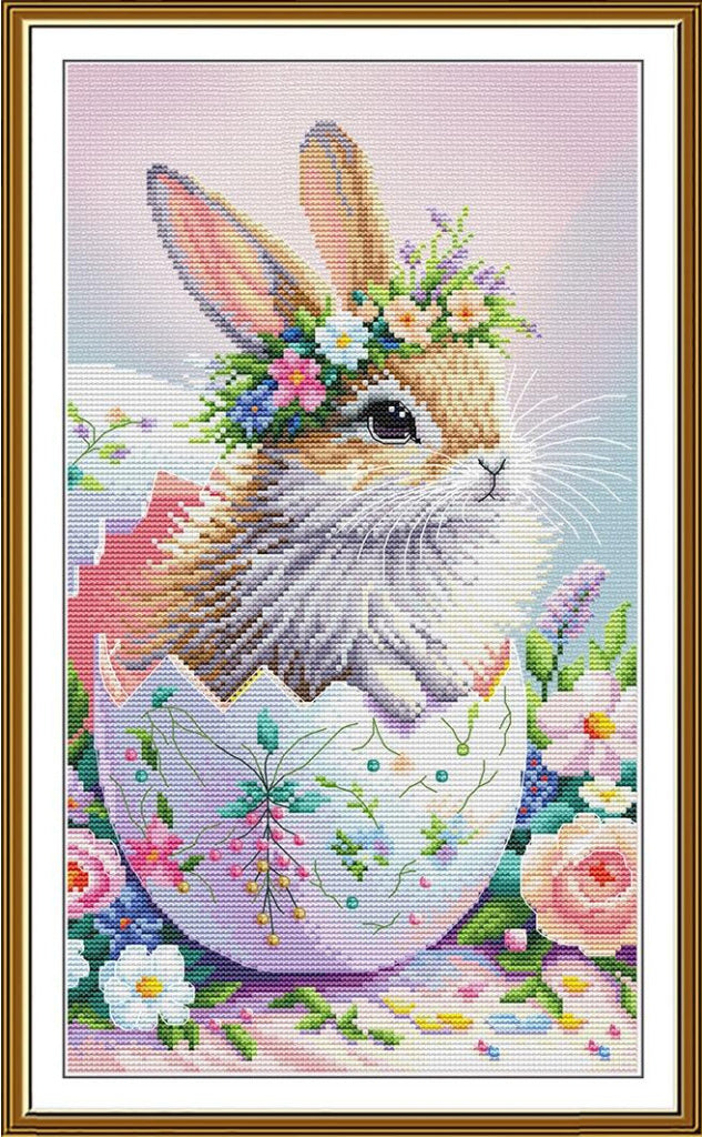 A framed cross-stitch or needlework-style artwork showing a fluffy white and tan bunny with a floral crown emerging from a decorated pastel egg, surrounded by flowers on a soft pink background.