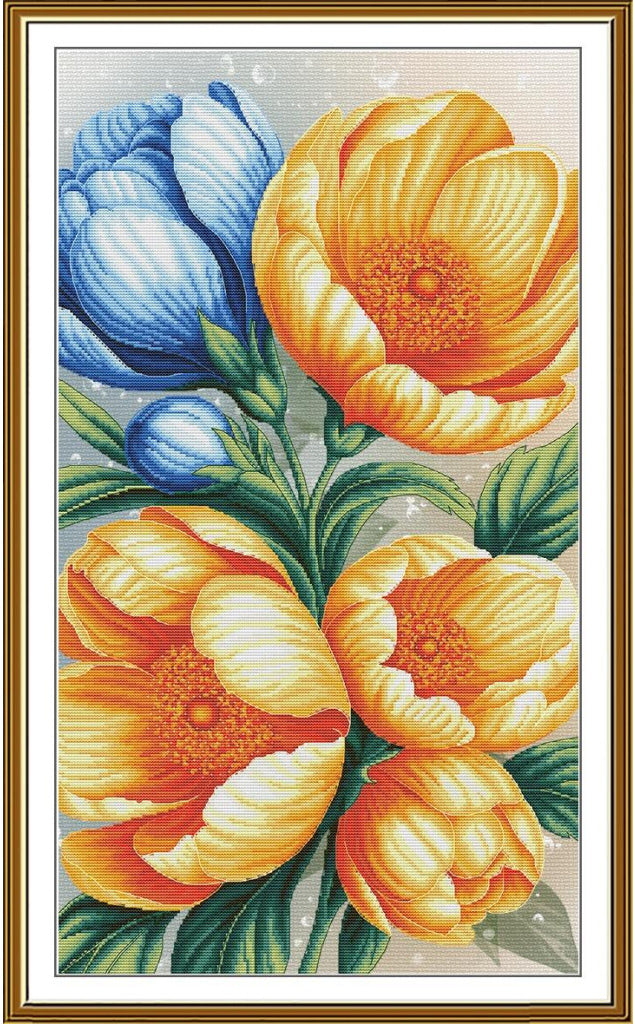 A framed floral needlepoint or cross-stitch style artwork showing large yellow-orange flowers, blue flower buds, and green leaves on a light background, surrounded by a gold-toned frame.