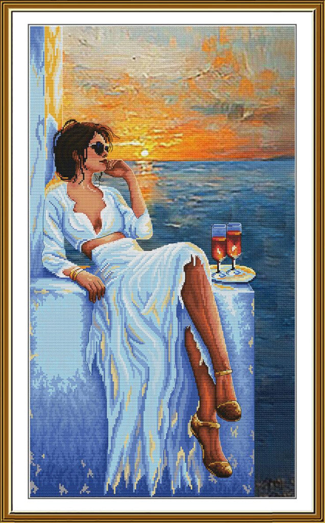 A framed canvas painting depicting a woman in a white dress sitting by the sea at sunset, with two glasses of red wine on a small table beside her. The artwork is shown within a gold-toned frame and white mat border.