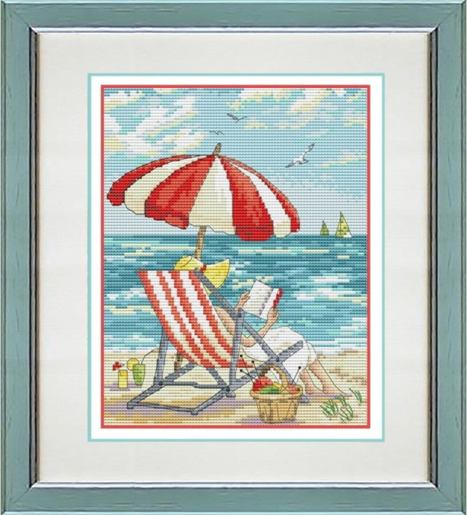 A framed cross-stitch artwork depicting a beach scene with a red and white striped umbrella, a matching striped deck chair, blue ocean water, and a pale sky with a few birds.