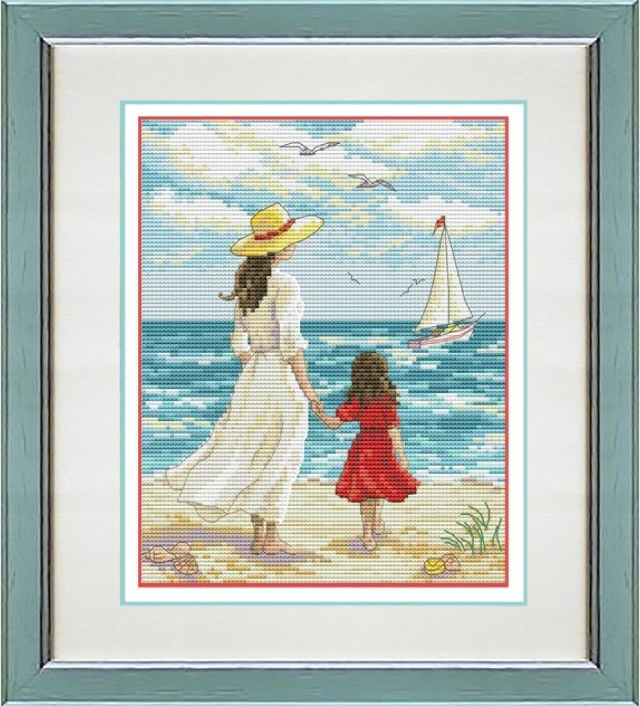 A framed cross-stitch or needlework artwork showing a woman in a white dress and yellow hat holding hands with a child in a red dress on a beach, with a sailboat and blue ocean in the background. The piece is mounted in a white mat and a muted green frame.
