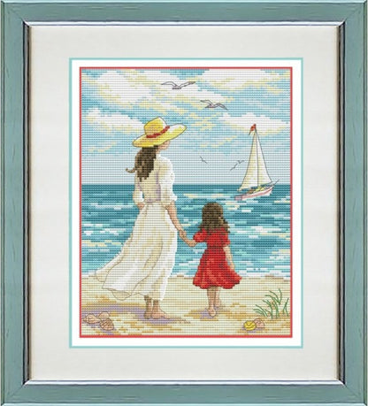 A framed cross-stitch or needlework artwork showing a woman in a white dress and yellow hat holding hands with a child in a red dress on a beach, with a sailboat and blue ocean in the background. The piece is mounted in a white mat and a muted green frame.