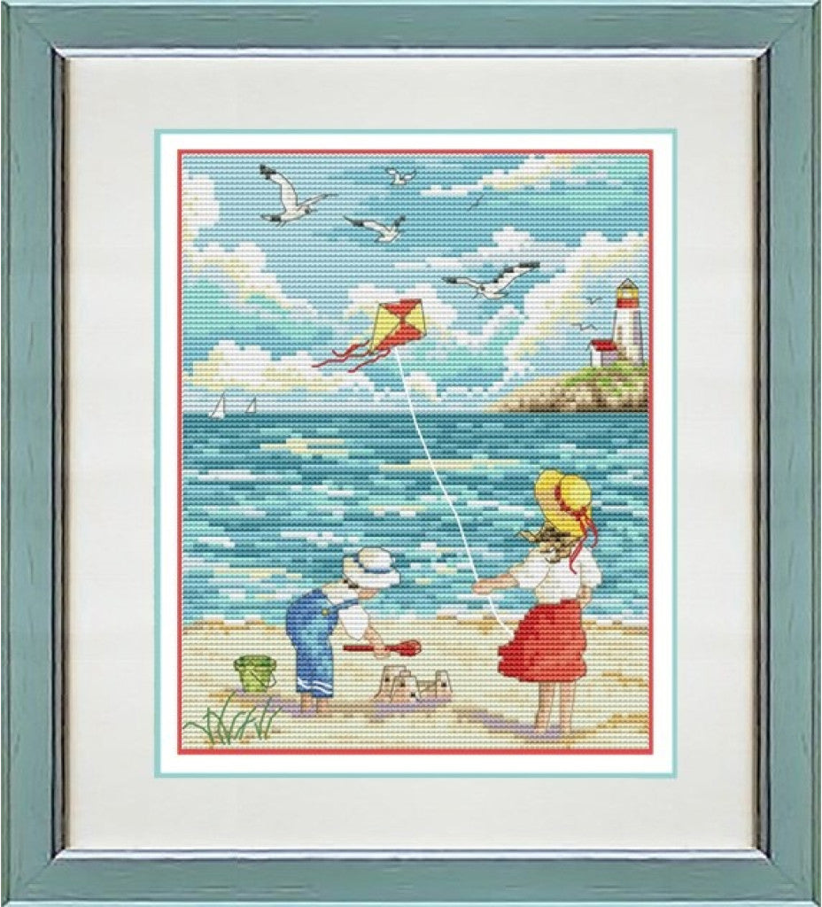 A framed cross-stitch or needlework artwork depicting a beach scene with two children flying a red and yellow kite, seagulls in the sky, a lighthouse, and blue water. The piece is matted and presented in a square gray frame.