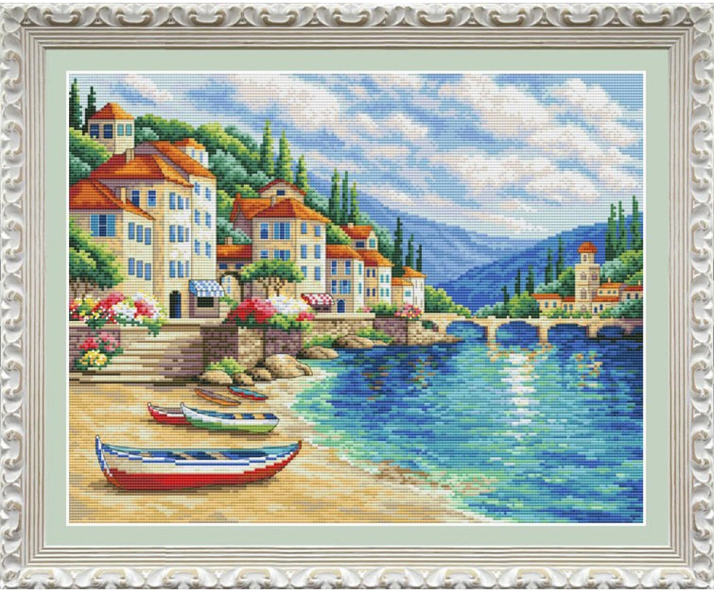A framed cross-stitch or needlepoint art piece depicting a coastal village scene with blue water, boats on a sandy shore, colorful houses with red-orange roofs, mountains in the distance, and a white ornate frame.